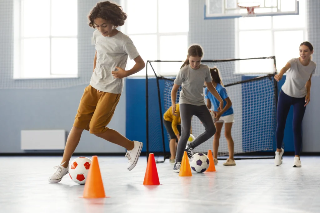 Grupo de estudiantes jugando fútbol en un gimnasio con conos, ilustrando cómo la actividad física en el aula potencia el rendimiento académico